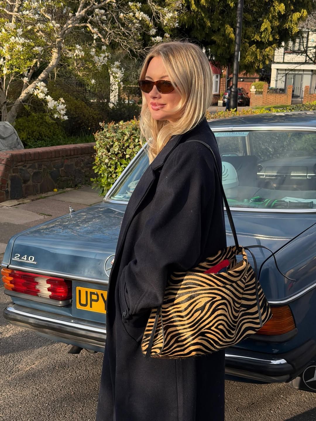 Blonde woman in black coat with zebra print bag poses beside a vintage Mercedes 240 on a suburban street.