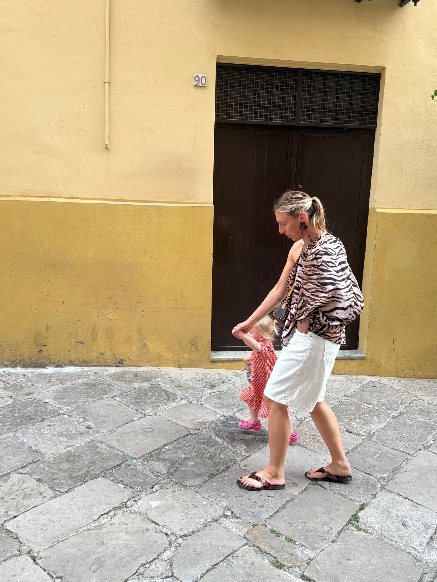 Woman in tiger print top and white skirt walking on cobblestone street past yellow building with dark wooden door.