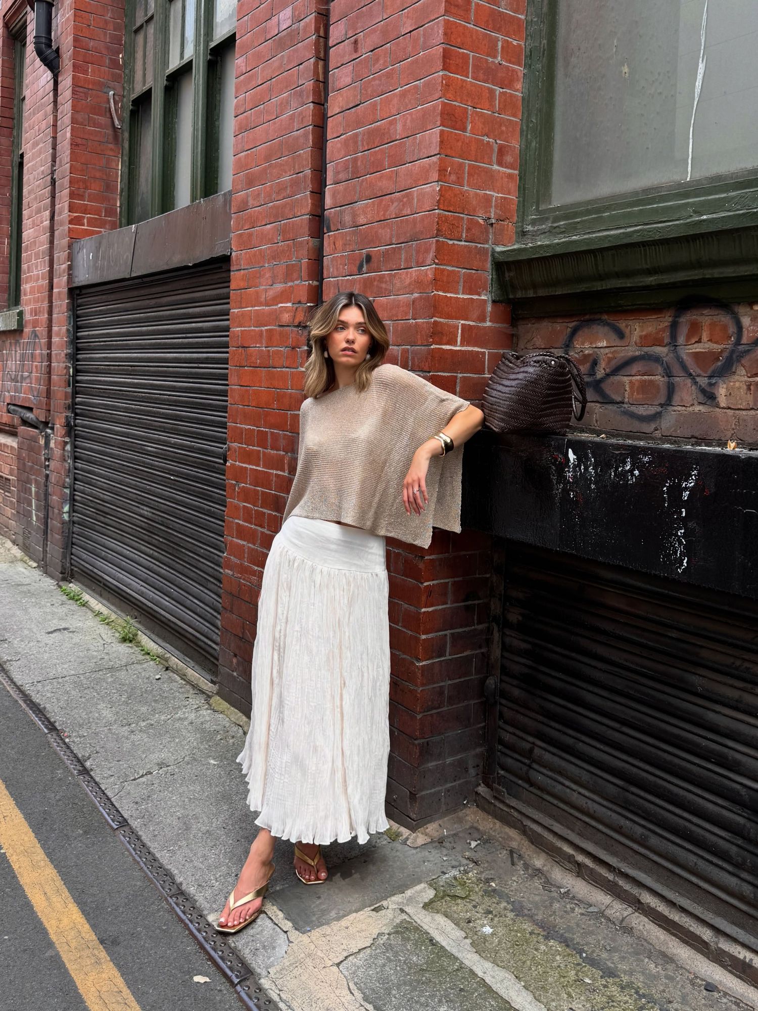 Woman in beige knit top and white pleated midi skirt posing in an urban alleyway with red brick walls.