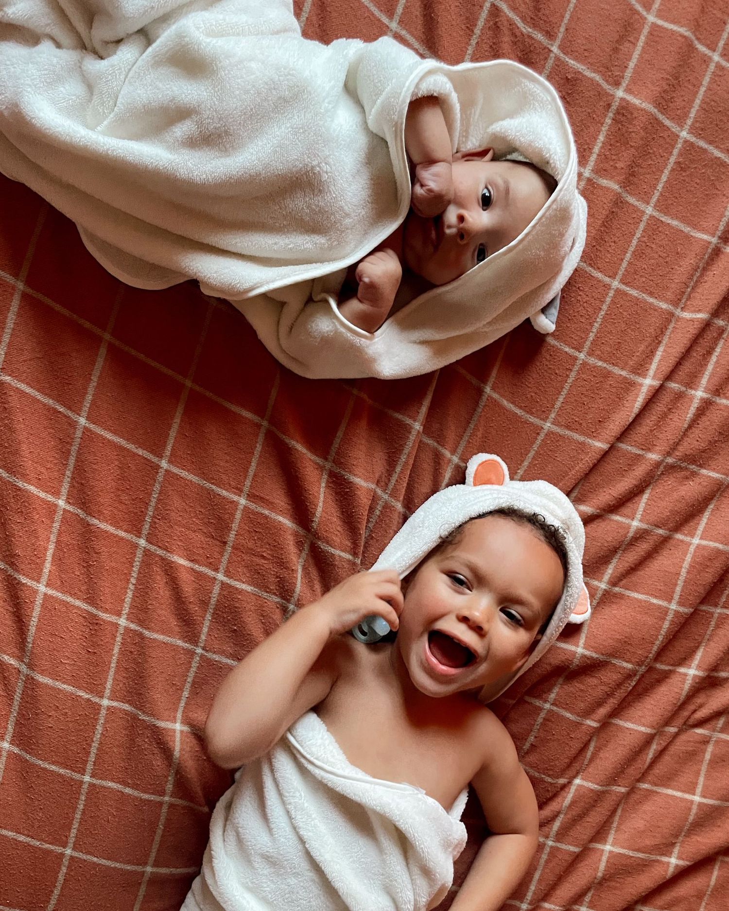 A baby in white towel lies on orange bedding while laughing and playing with outstretched arms.