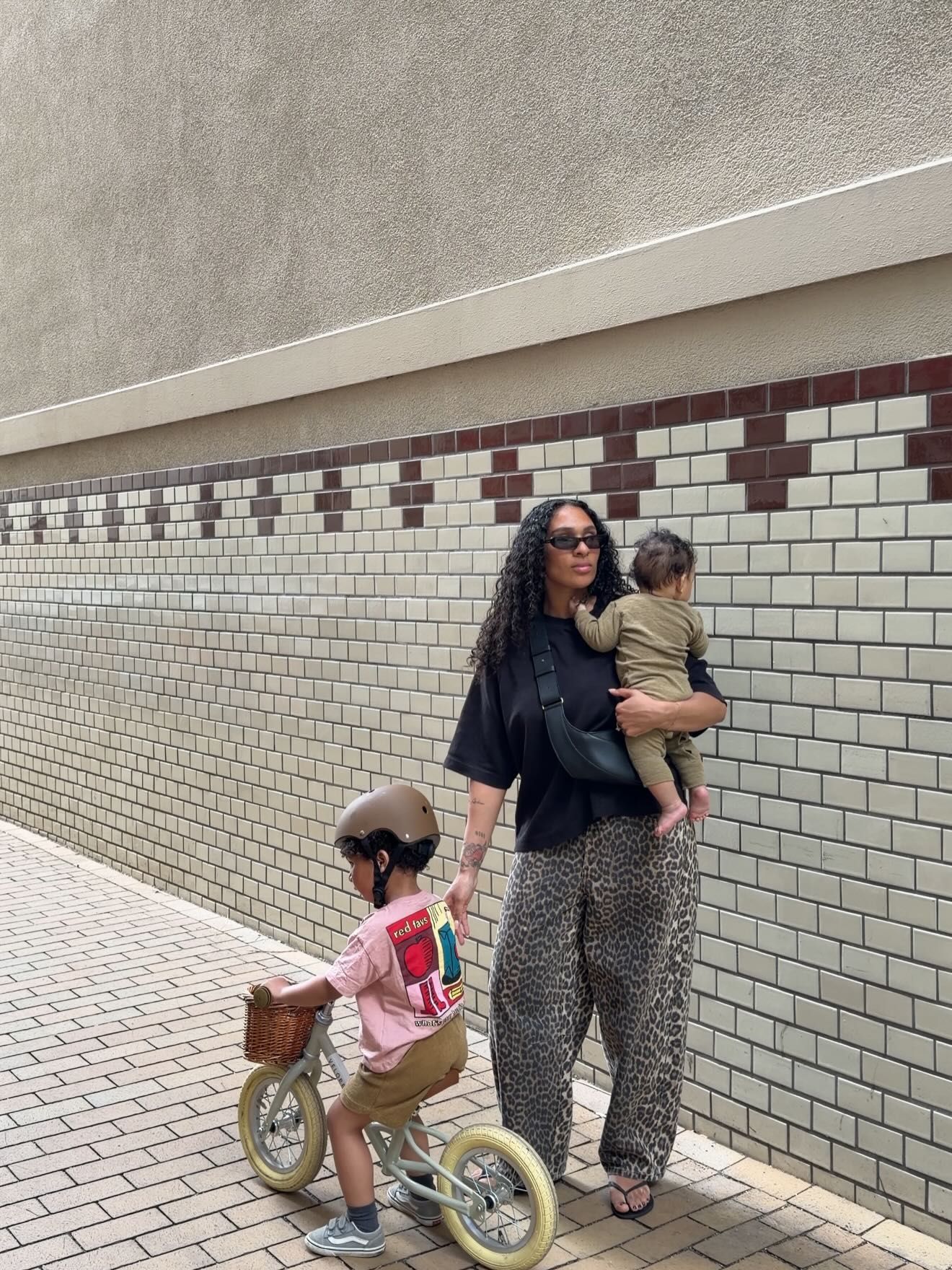 Woman holding toddler in leopard print pants stands by tiled wall while child rides balance bike on sidewalk.
