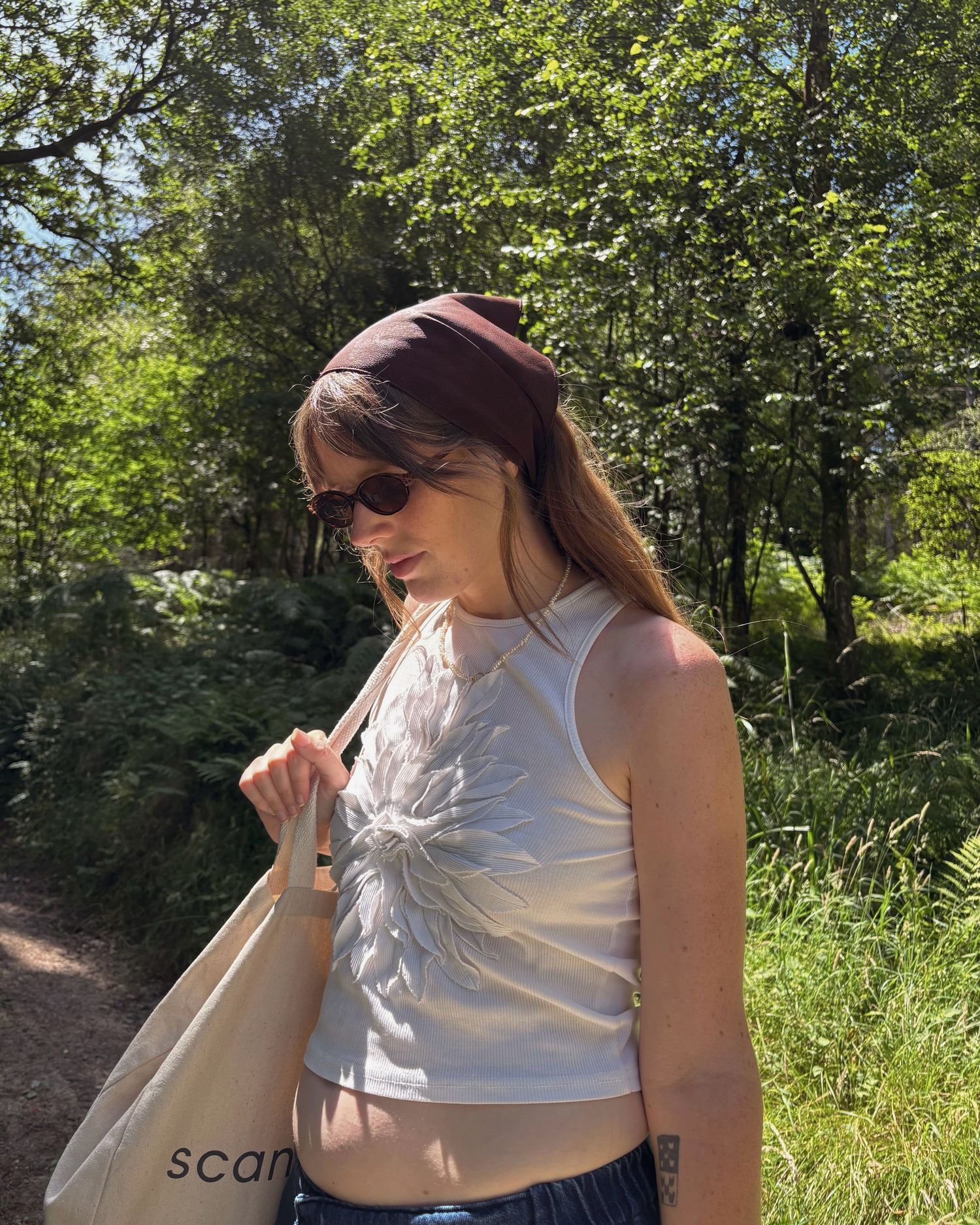 Young woman in white crop top, burgundy headband, and sunglasses carrying a tote bag outdoors on a sunny day.