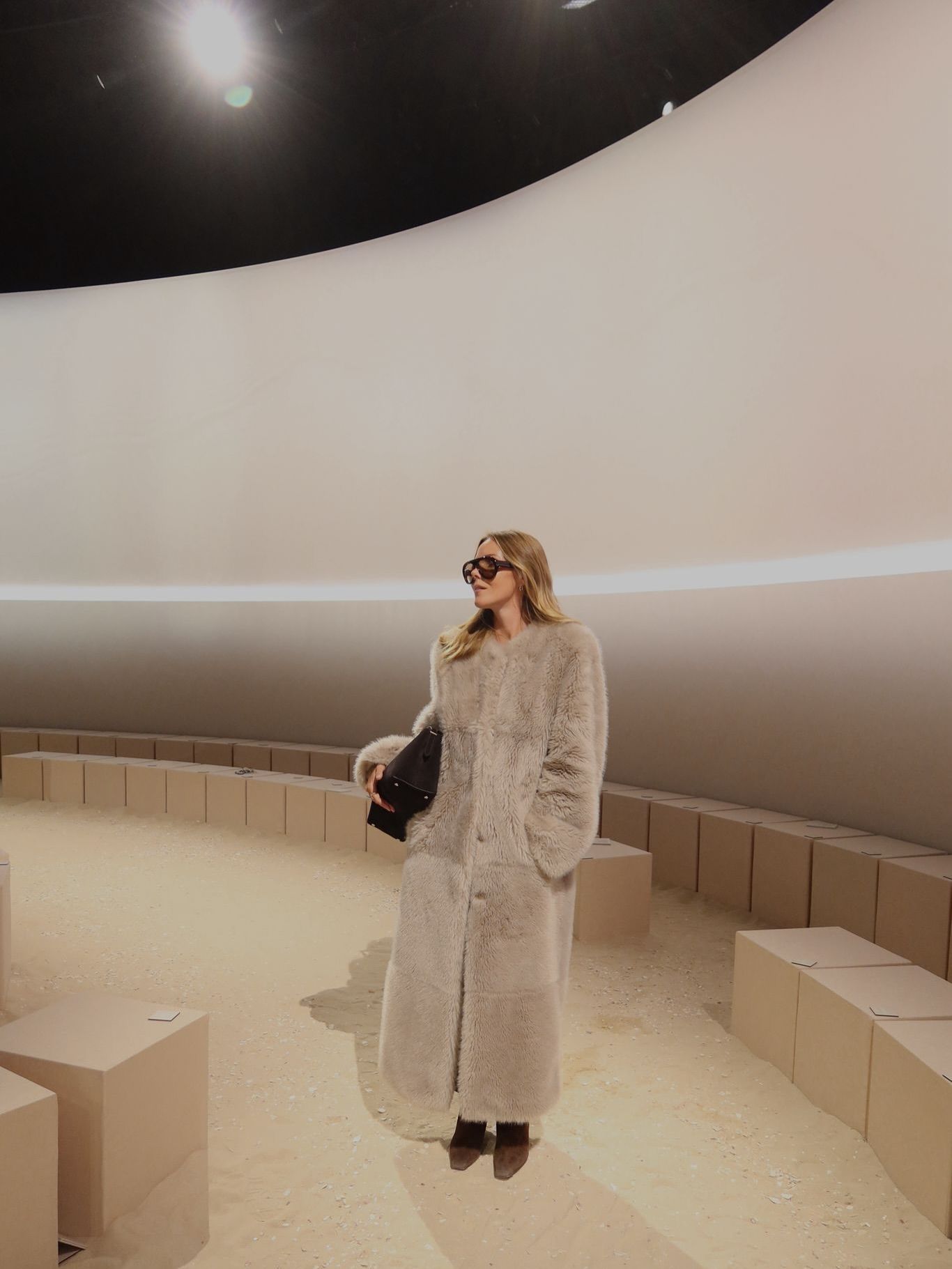 Woman in long cream shearling coat holding black bag, standing in minimalist curved interior space.