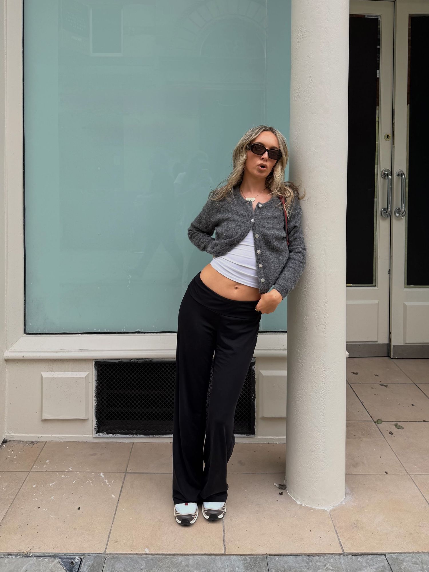 Woman in gray cardigan, white crop top, black wide-leg pants, and sneakers posing against a teal wall.