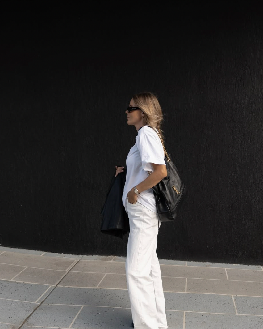 A person in white clothing walks past a dark wall while casting a shadow on the ground.