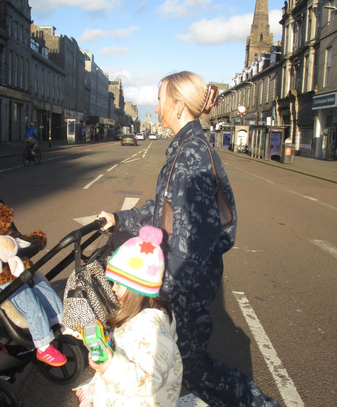 Blonde woman pushing a stroller with a child wearing a rainbow hat on a sunny urban street.