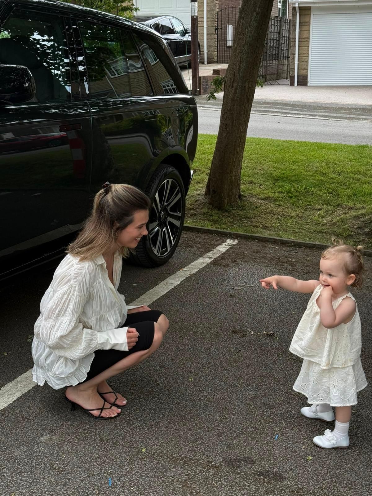 A woman crouches down smiling at a toddler in a white dress pointing at her in a parking lot.