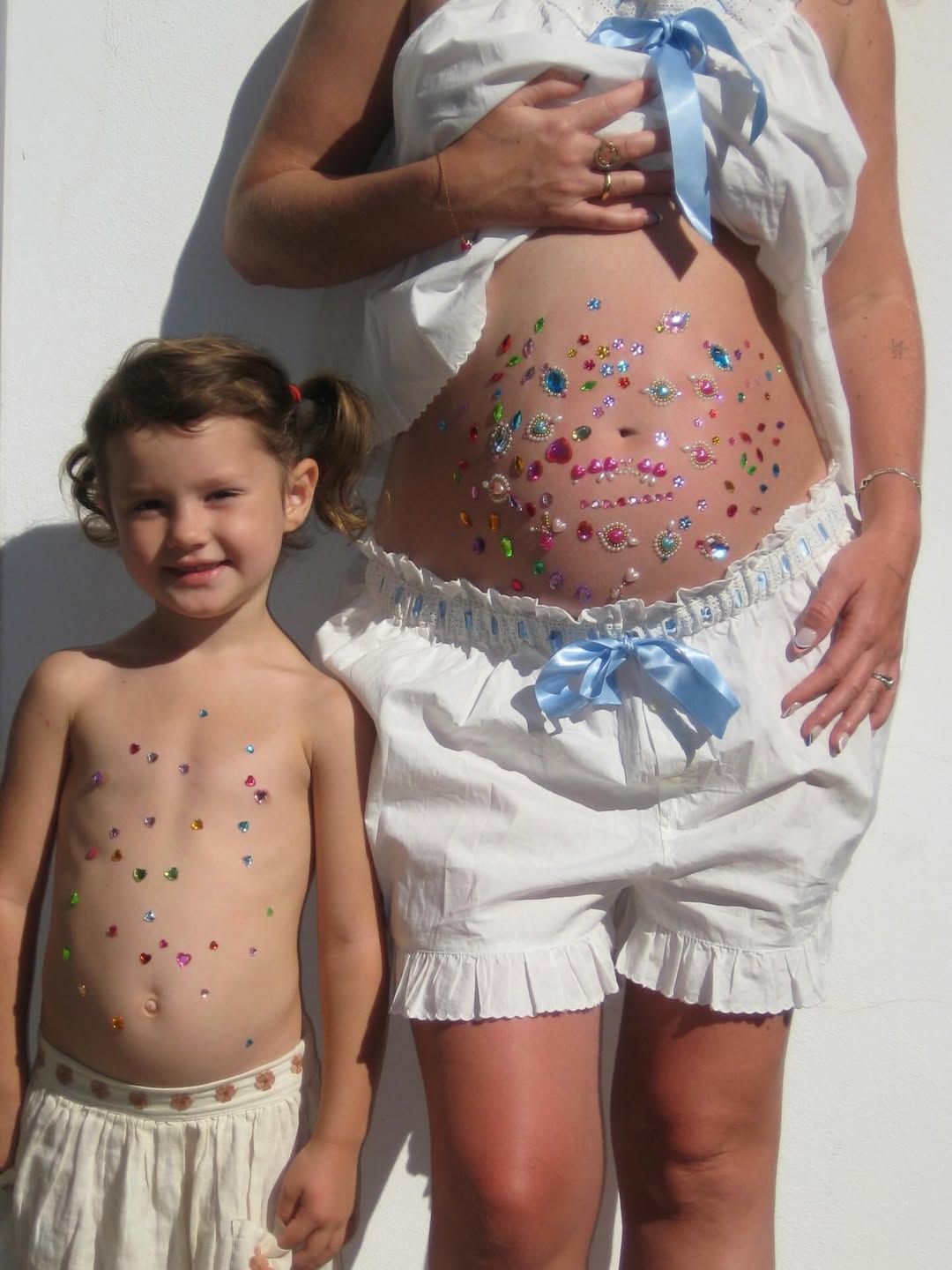Pregnant woman and young child with colorful gem stickers on their bellies, wearing matching white outfits outdoors.