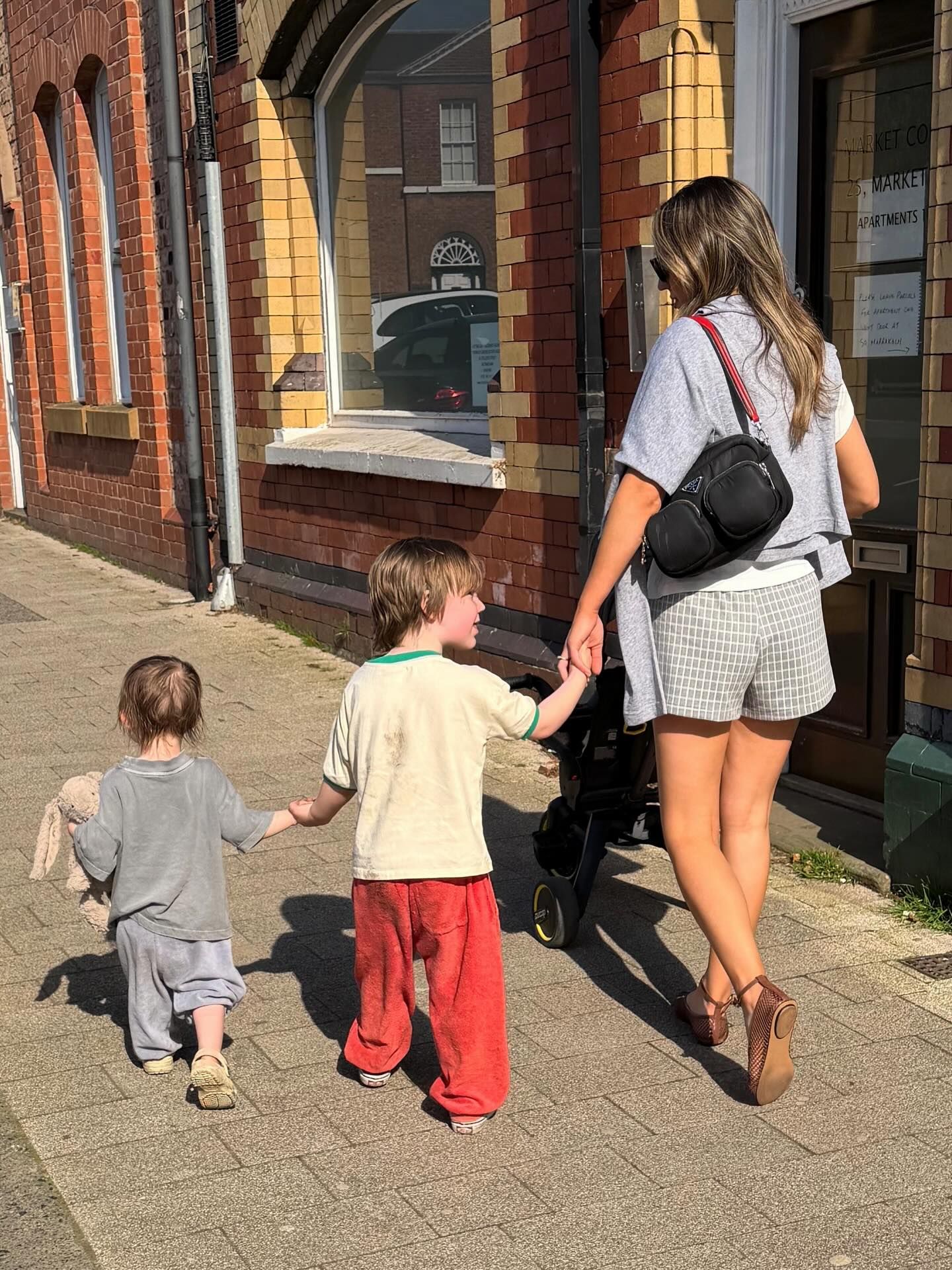 A woman walks hand-in-hand with two young children along a sunny brick building sidewalk, carrying a black crossbody bag.