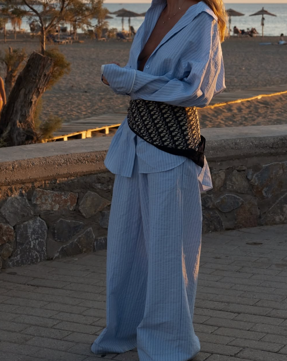 A person wearing a light blue oversized suit and polka dot dress poses on a stone wall at sunset by the beach.