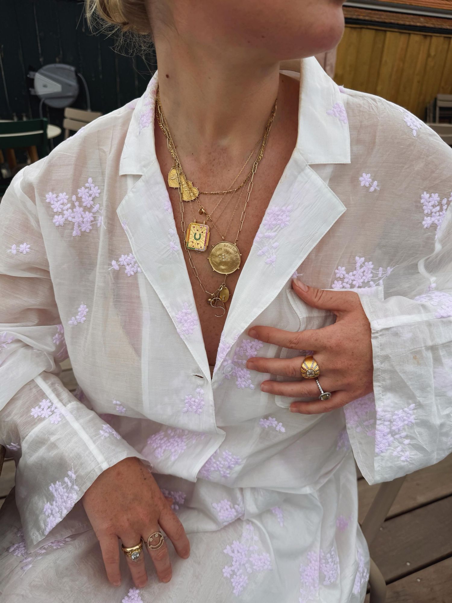 Woman wearing white floral sheer shirt with layered gold necklaces and gold rings, hand placed on chest.