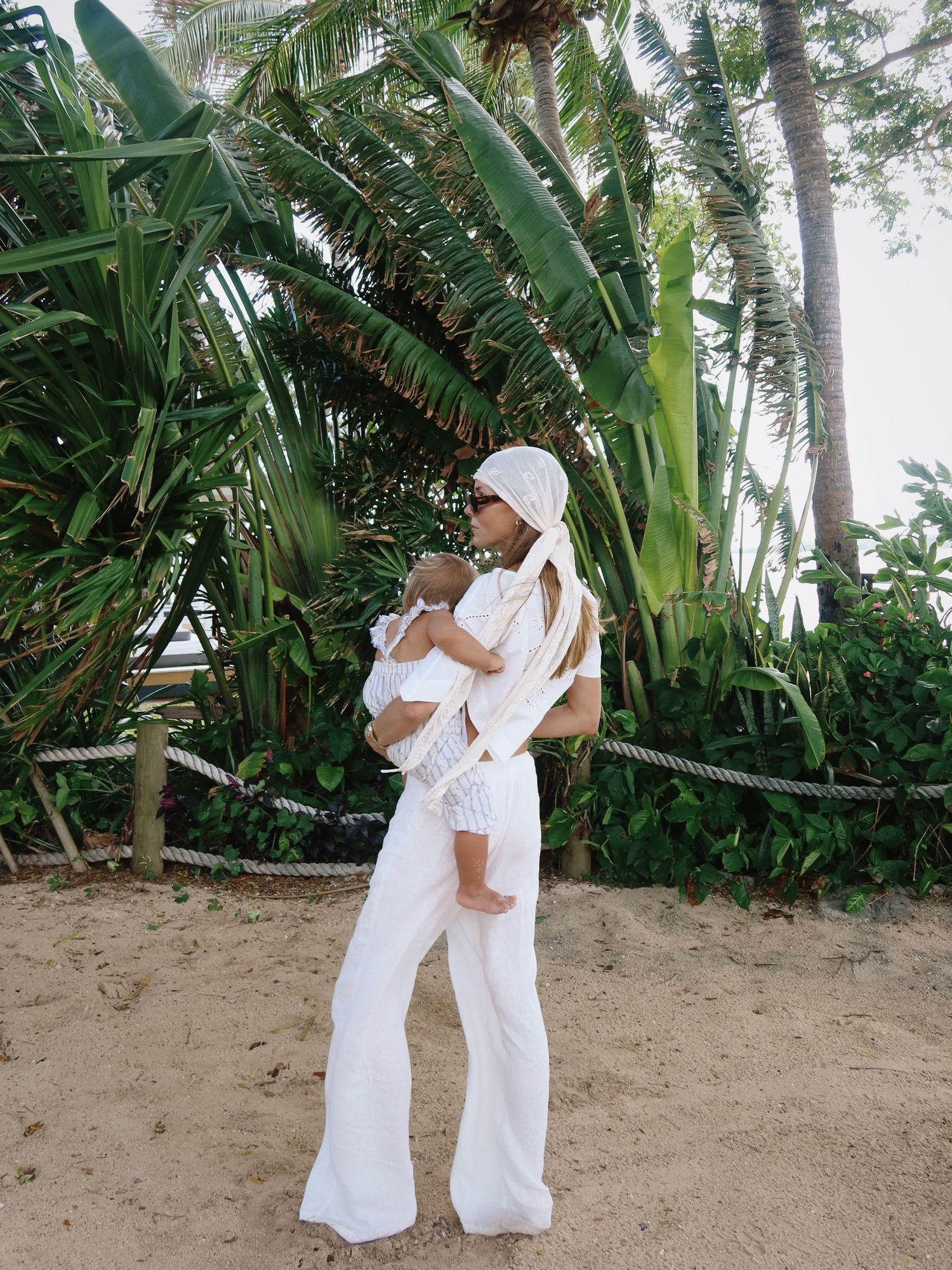 Woman in white outfit holding baby on sandy beach with tropical palm trees in background.