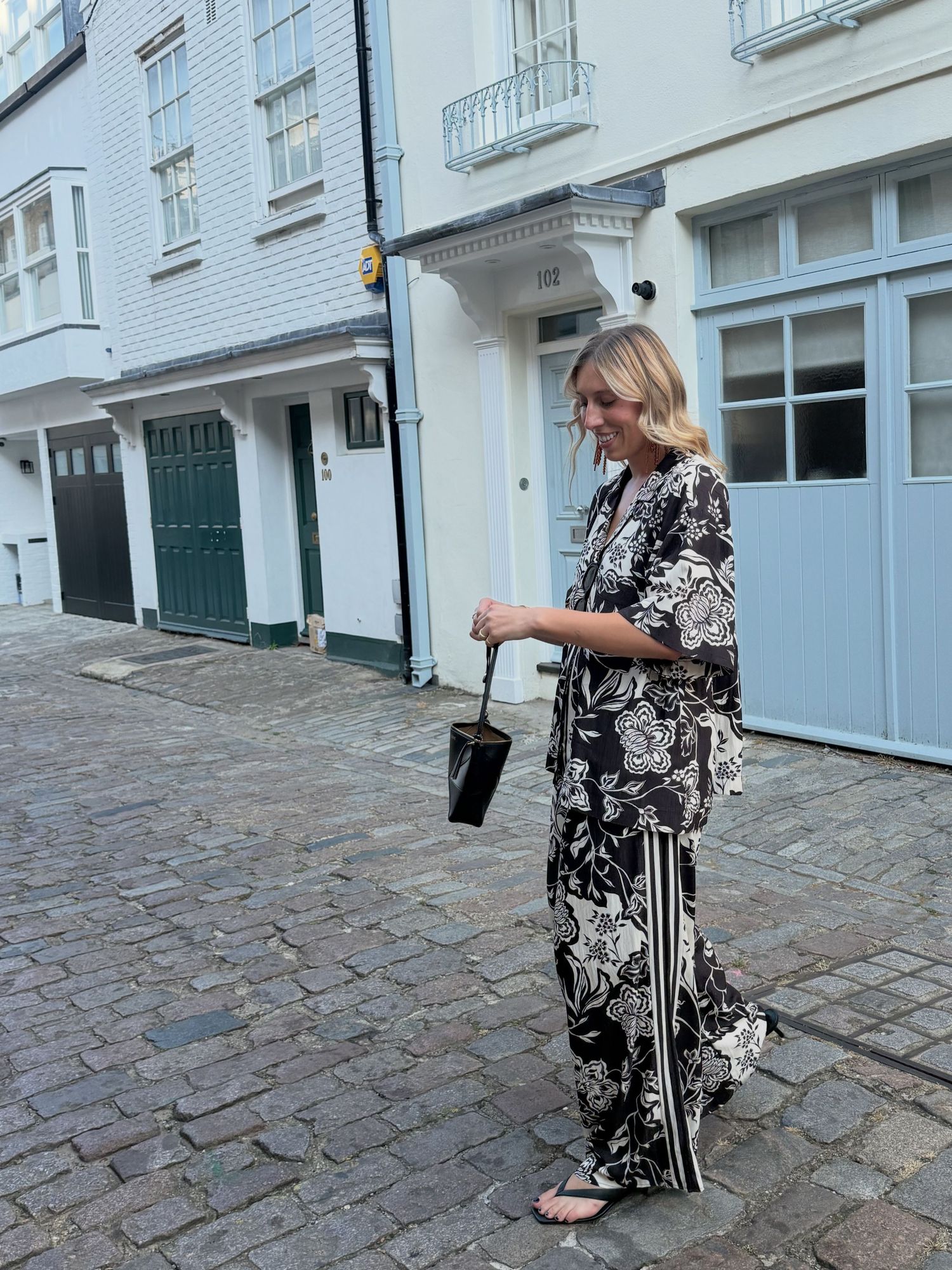 Woman in black floral wide-leg pants and matching top walks on cobblestone mews street carrying small black bag.