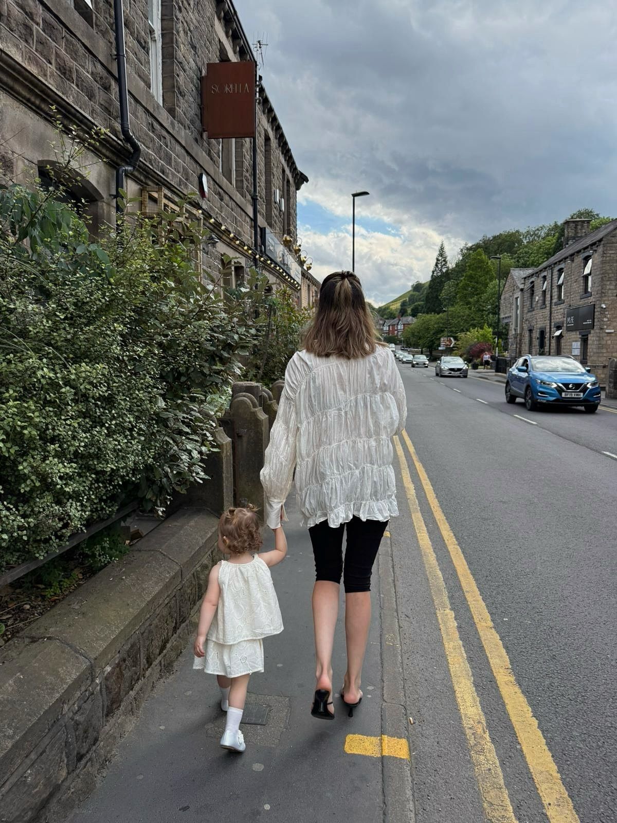 A woman and young girl holding hands walking along a quiet street beside a stone building in a UK village.
