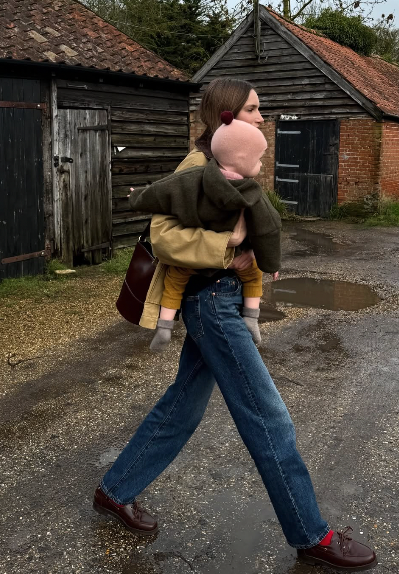 Someone in wide-leg jeans and a brown jacket walks past rustic wooden barns on a rural path.