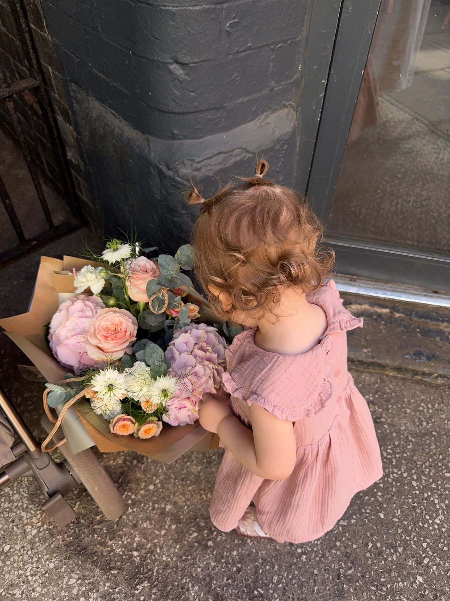 Toddler girl in pink dress smelling fresh flower bouquet with peonies and roses outside a shop.