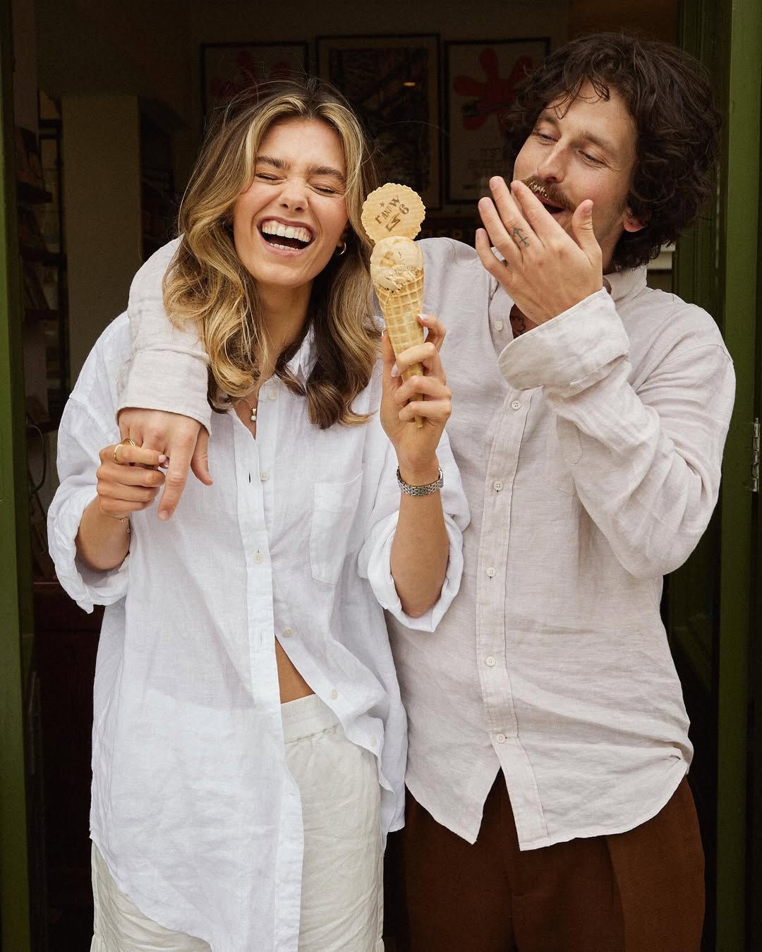Laughing couple in white linen outfits sharing an ice cream cone outside a green doorway.