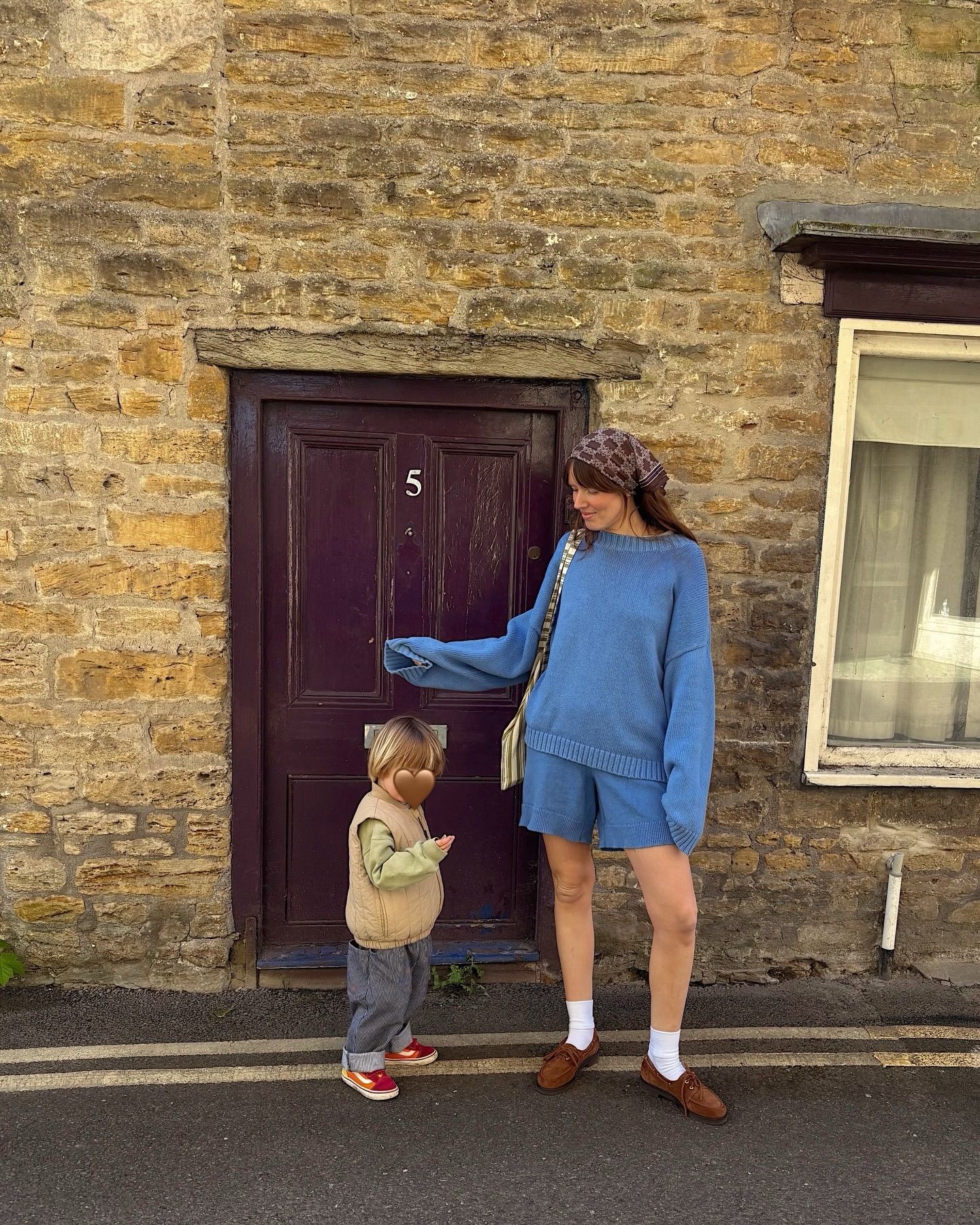 Woman in blue matching set stands with toddler by stone cottage door numbered 5.
