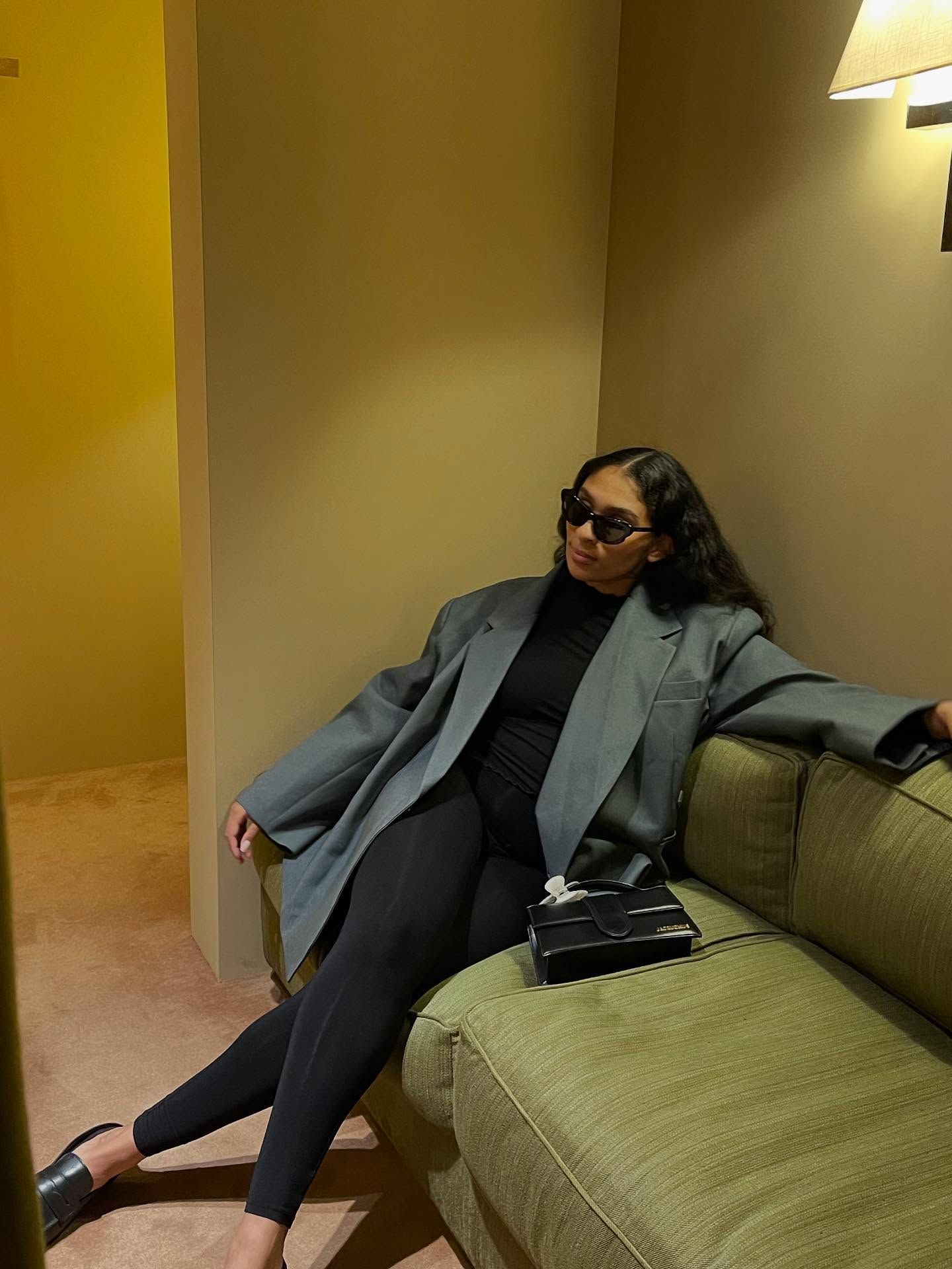 A stylish woman in an oversized gray blazer and black outfit relaxes on a green sofa wearing sunglasses.