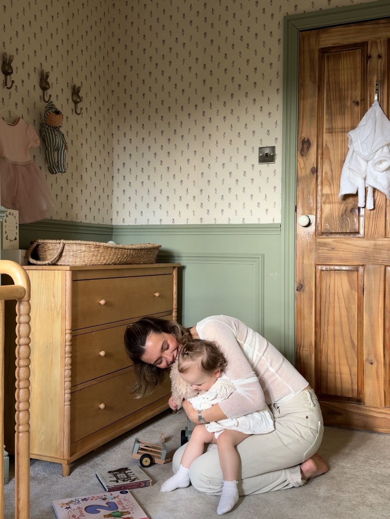 Mother kneeling on nursery floor hugging toddler near wooden dresser with green wainscoting and floral wallpaper.