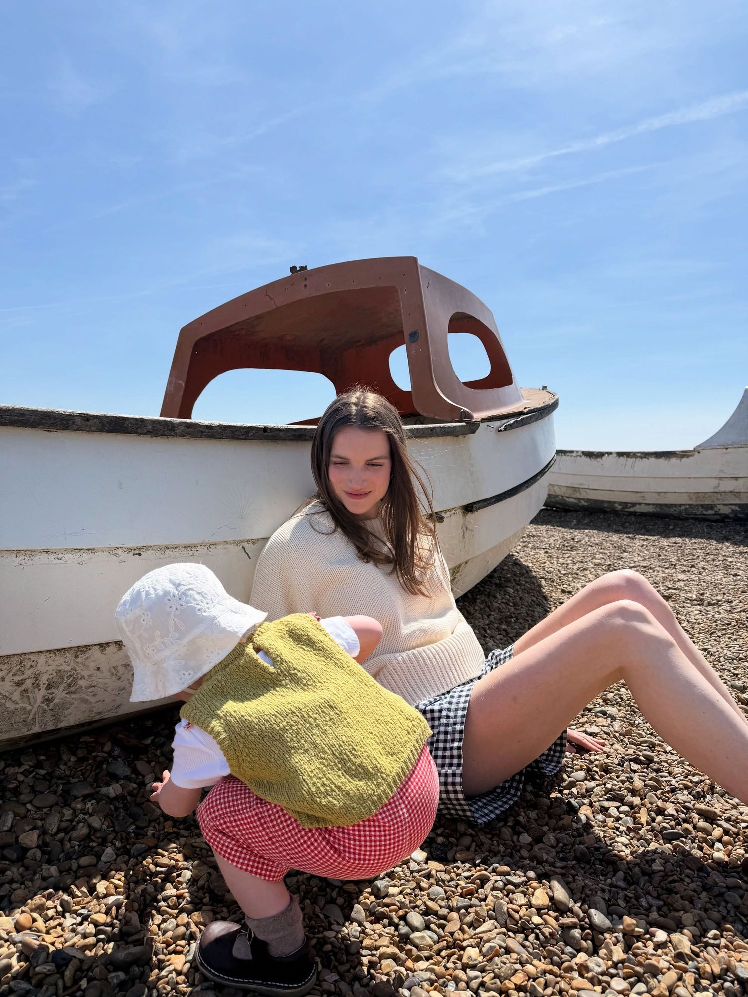 Young woman sitting on pebble beach leaning against a white boat, holding a green bag on a sunny day.