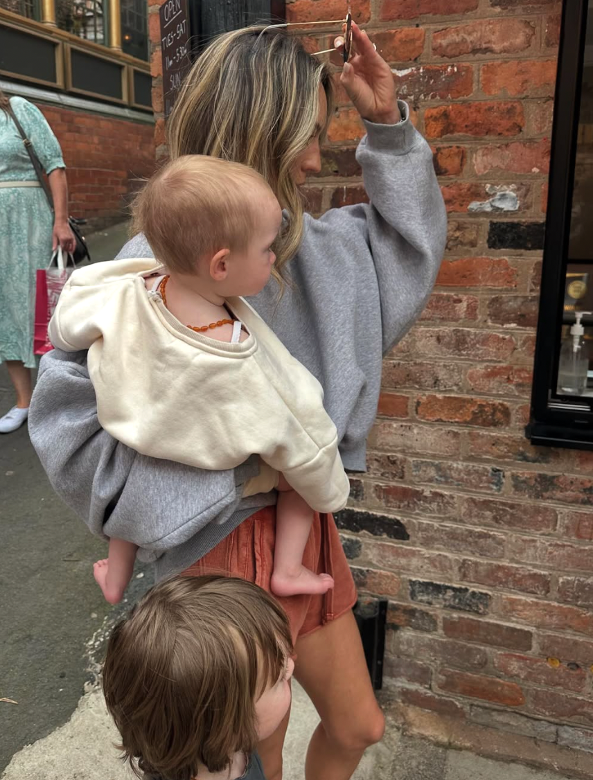 A group stands against a brick wall, with one wearing a gray sweatshirt and holding a small child in a beige top.