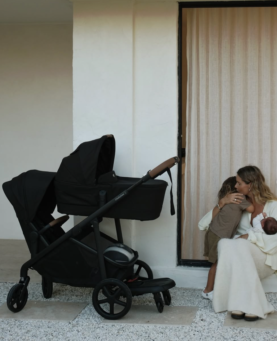 A black baby stroller parked next to a wall while someone sits on a white bench nearby.