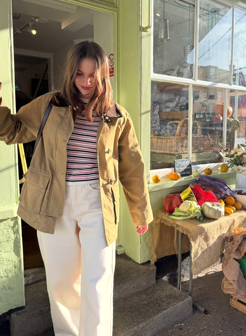 A person in a beige jacket and striped shirt poses against a storefront window in a casual outfit.
