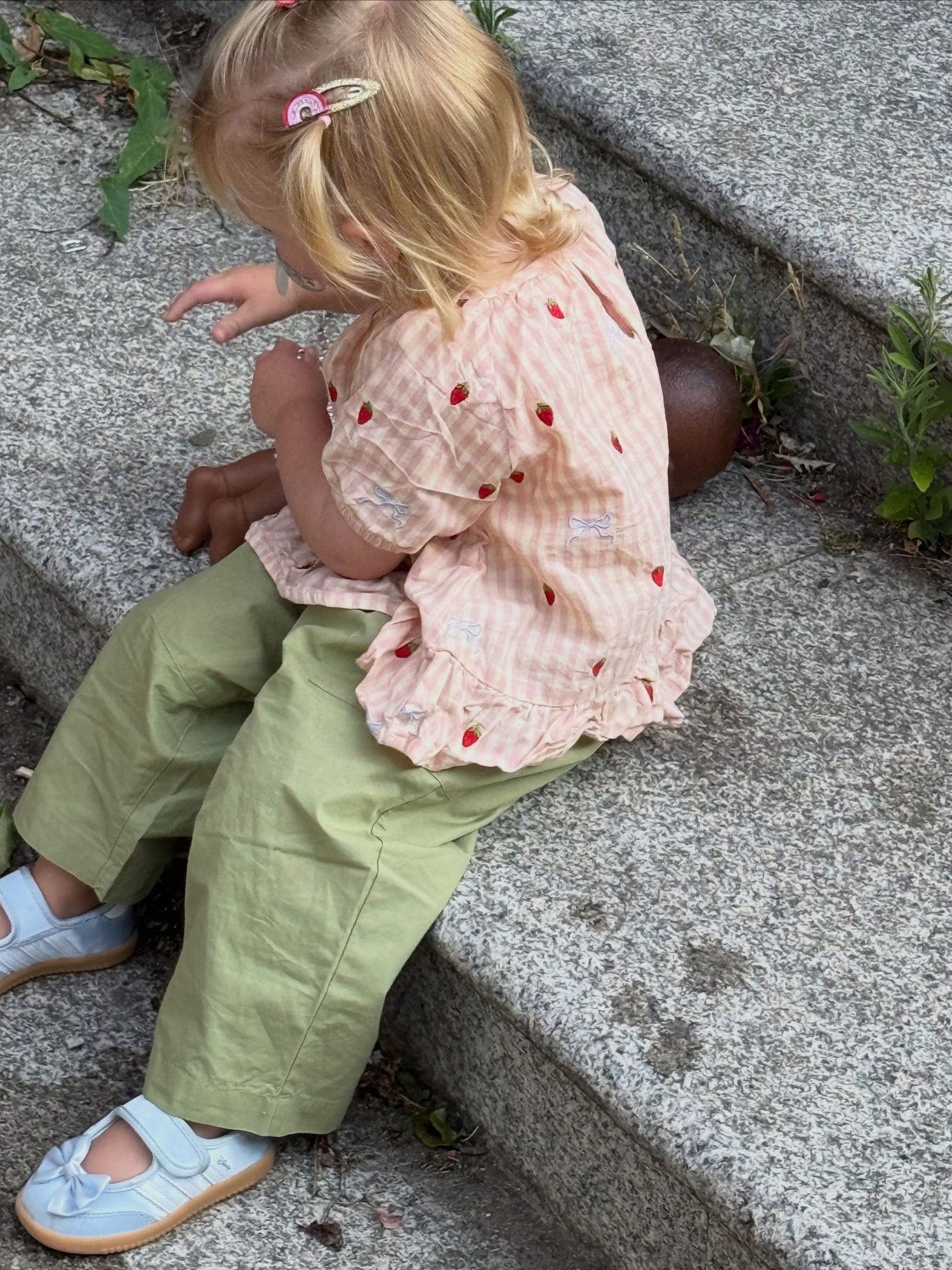 Toddler girl in green pants and strawberry print top sitting on stone steps, viewed from behind.