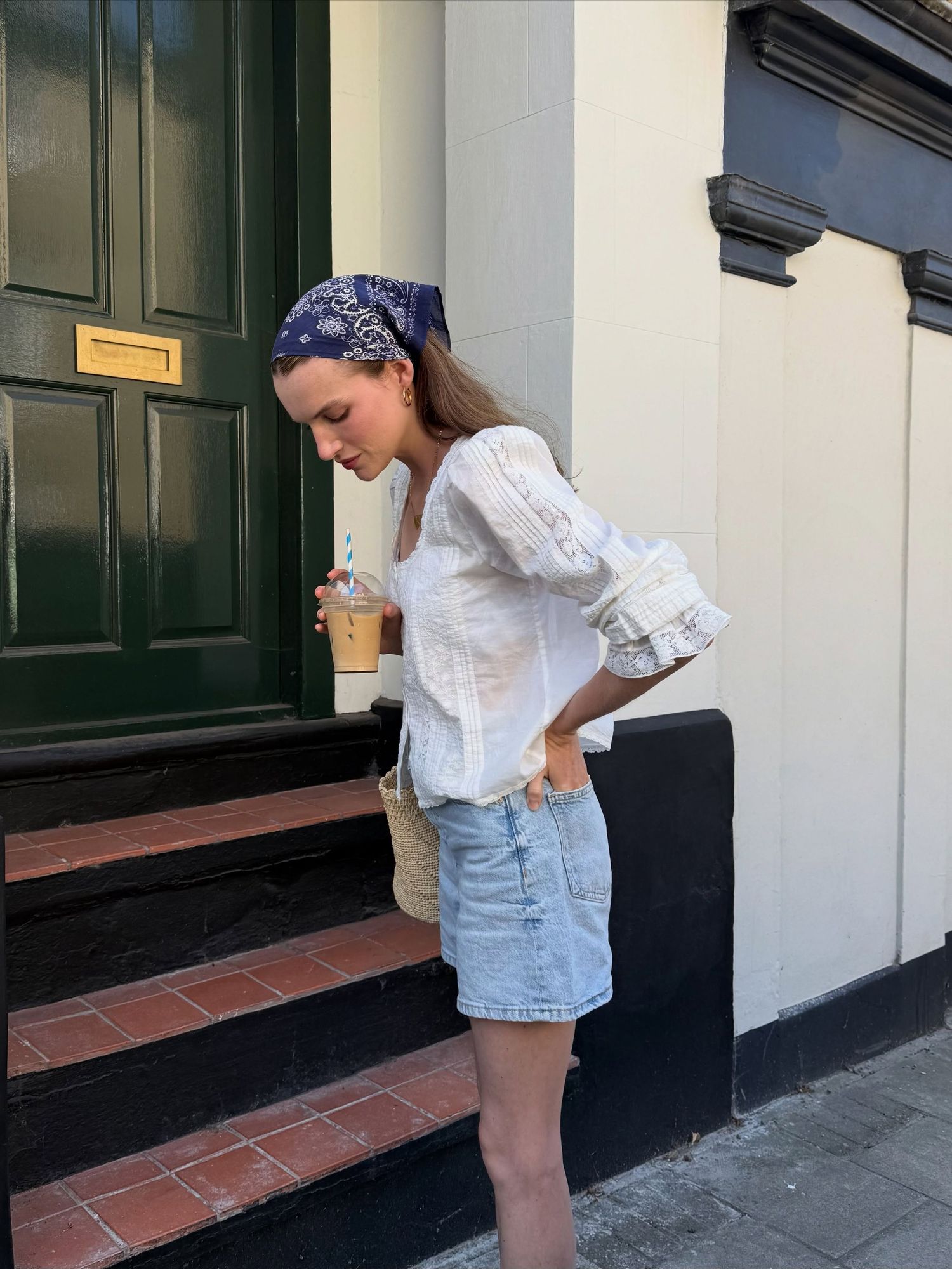 Woman in white blouse, denim skirt, and blue bandana headscarf holding iced coffee on steps by green door.