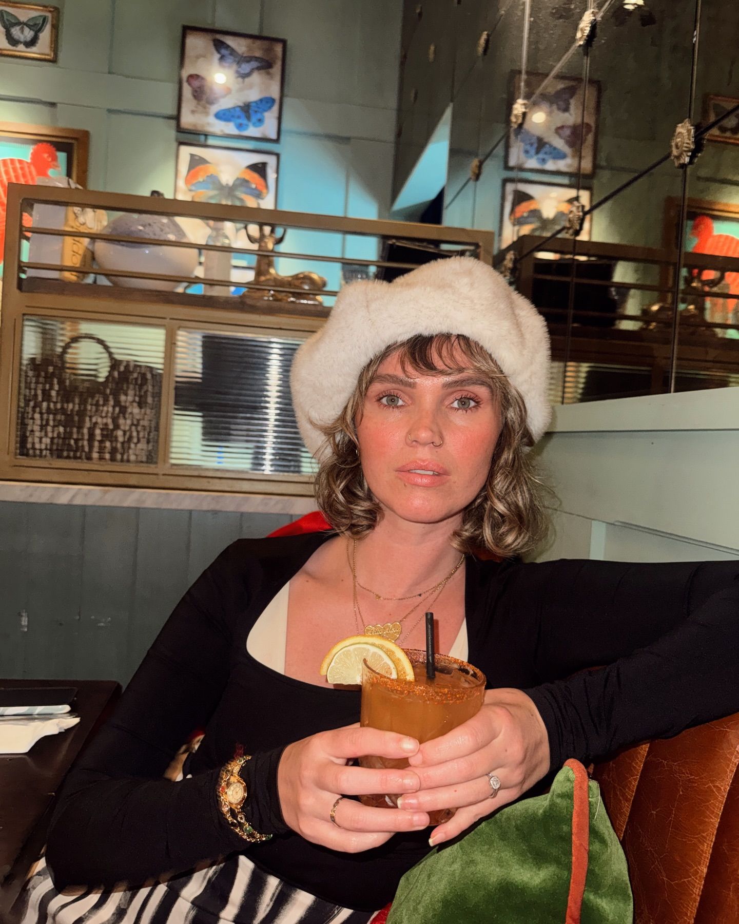 Woman in white fur hat holding a cocktail with lemon garnish at a stylish bar with butterfly wall art.