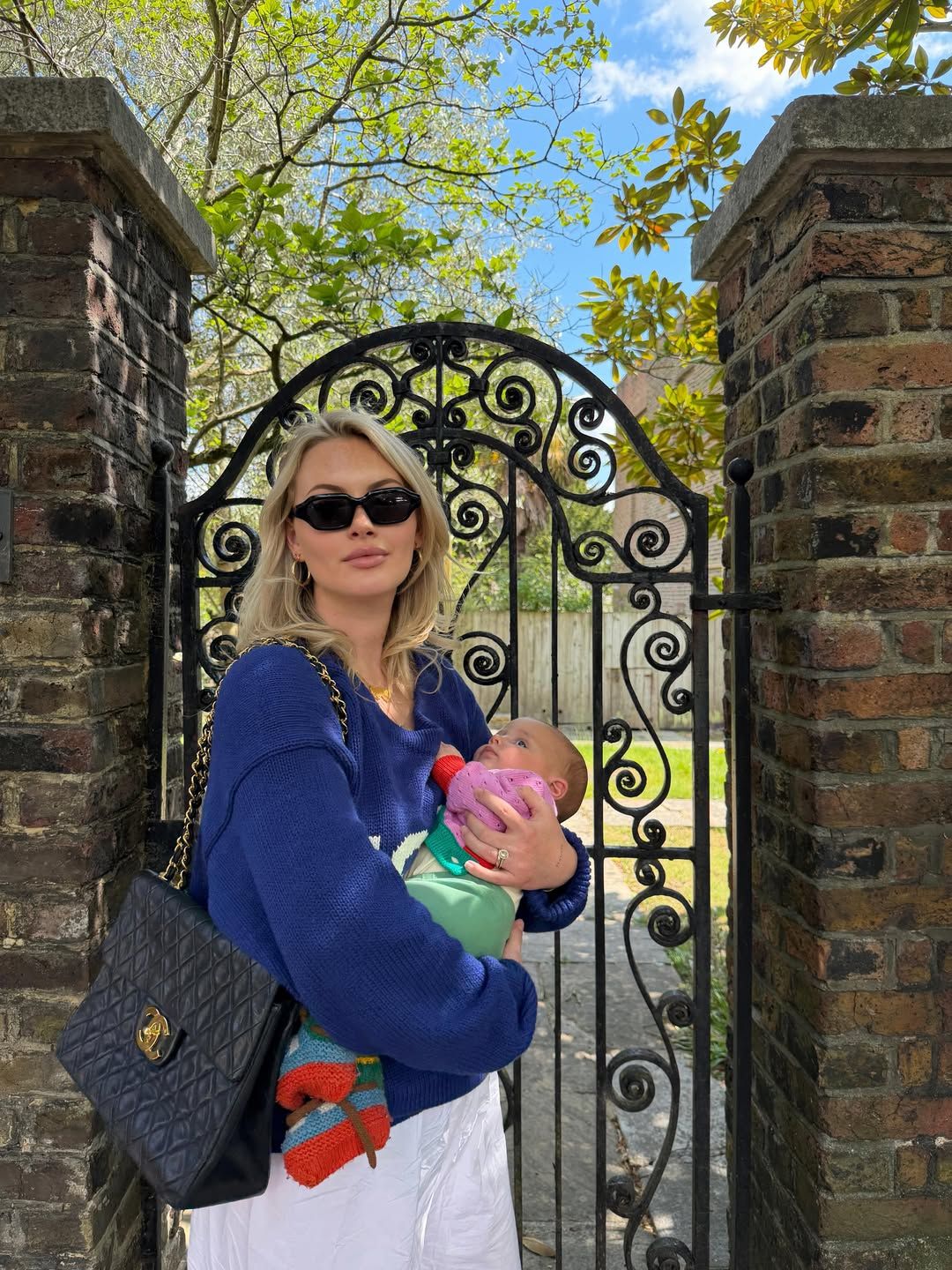 Woman in blue sweater holding a baby near ornate iron gate with brick pillars and lush green trees.
