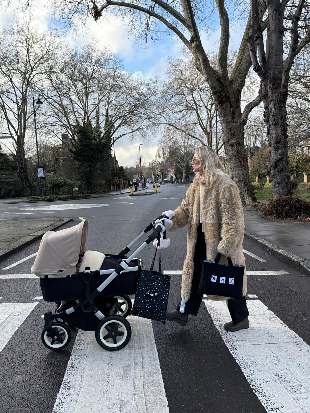 Woman in fur coat pushing a Bugaboo stroller across a zebra crossing on a tree-lined street.