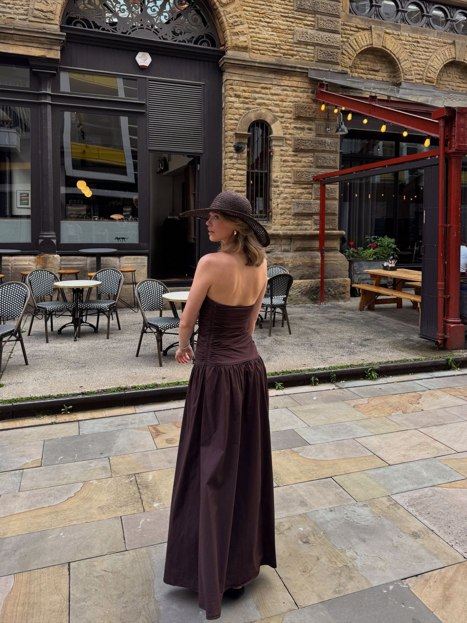 Woman in strapless dark brown maxi dress and matching hat poses on cobblestone street outside a bistro.