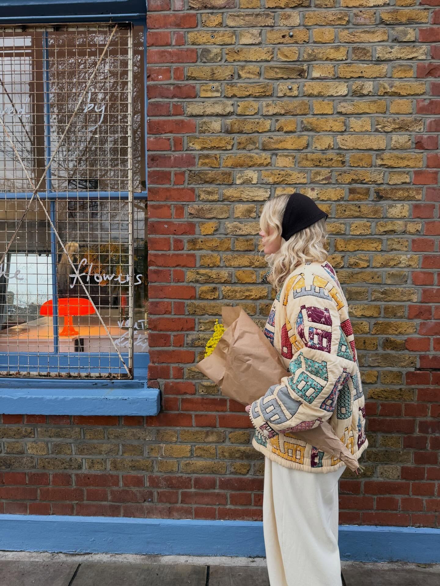 Woman in patterned jacket and black beanie holding flowers outside a brick wall shop.