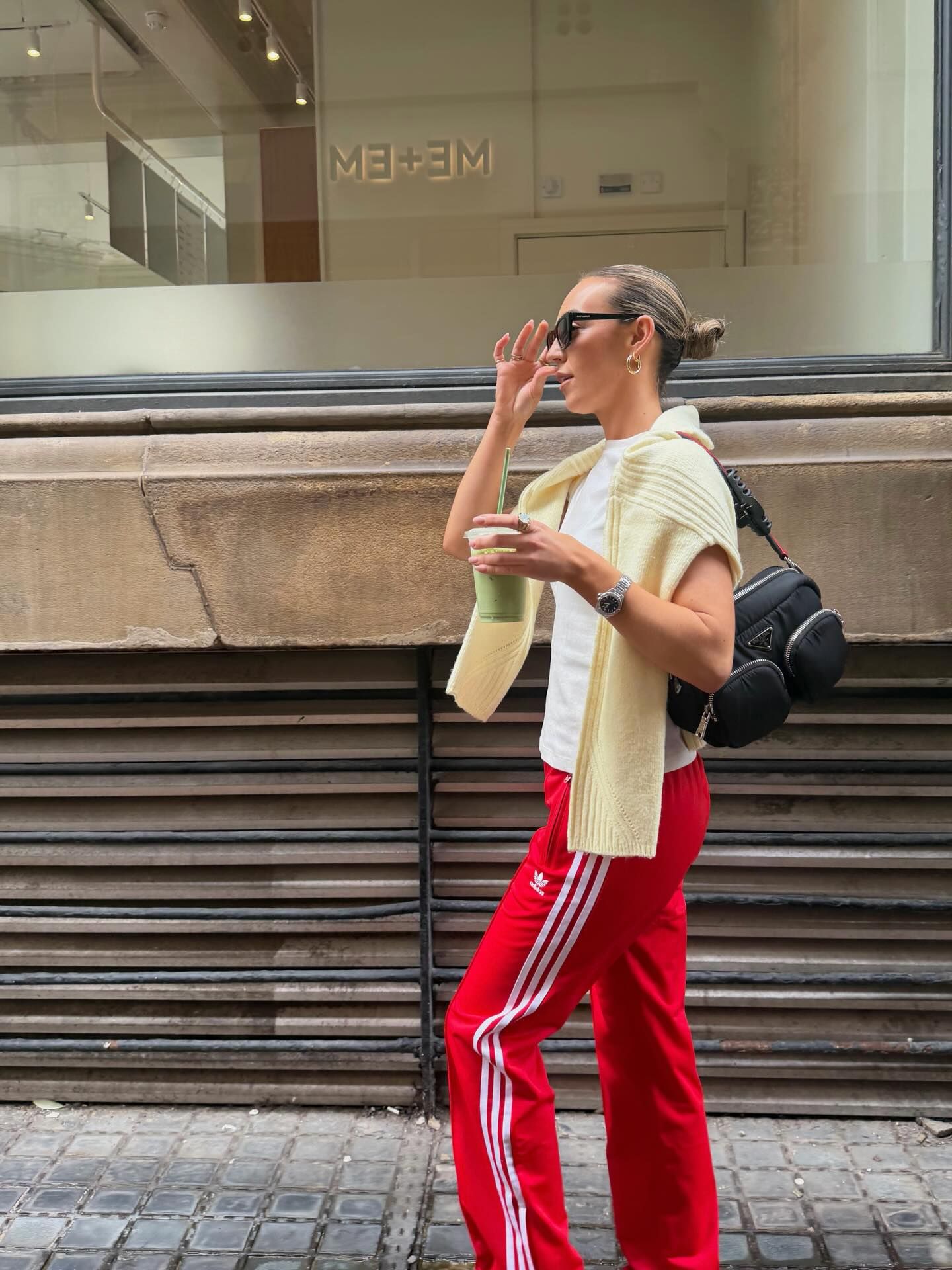 Woman in red Adidas track pants, cream knit vest, white tee, black bag, and sunglasses walking on a city street.