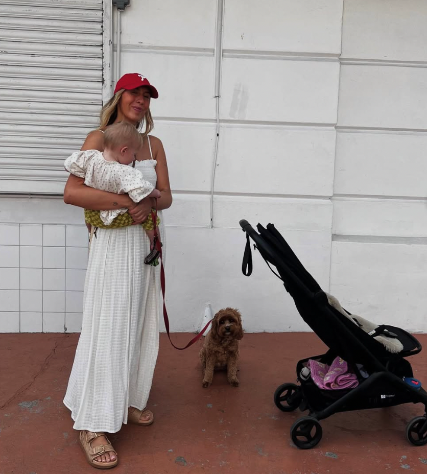 A fashionable person in a white maxi dress and red cap stands with a small brown dog next to a baby stroller.