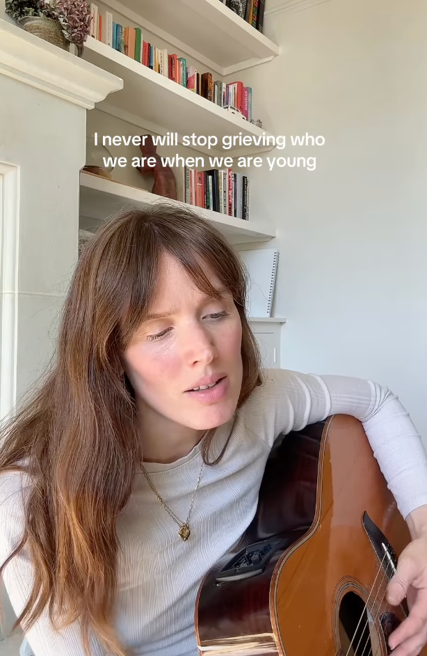 A person plays an acoustic guitar while sitting indoors with bookshelves visible in the background.