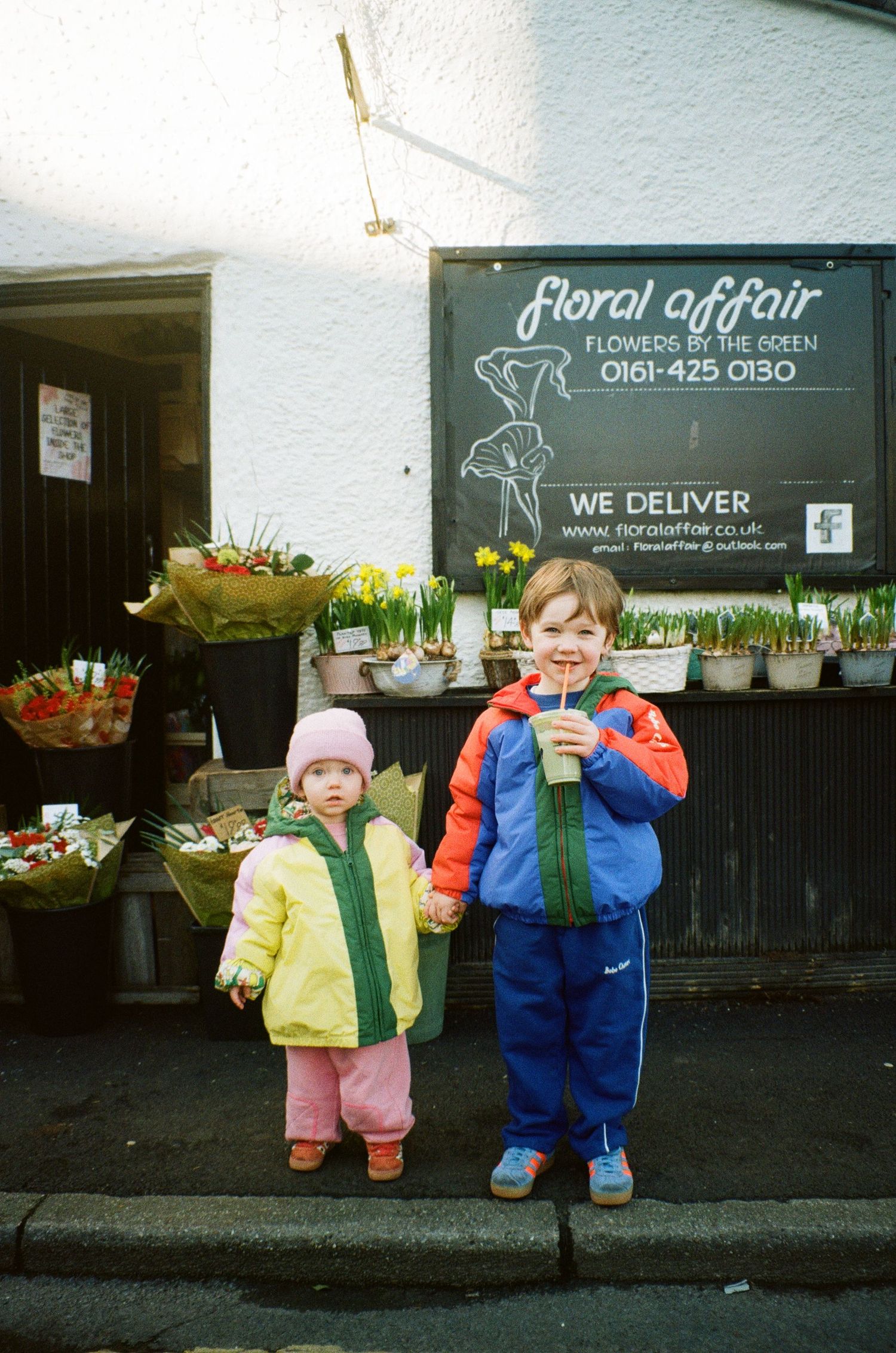 Two small children in puffy winter coats stand outside a flower shop called 'Floral Affair'.
