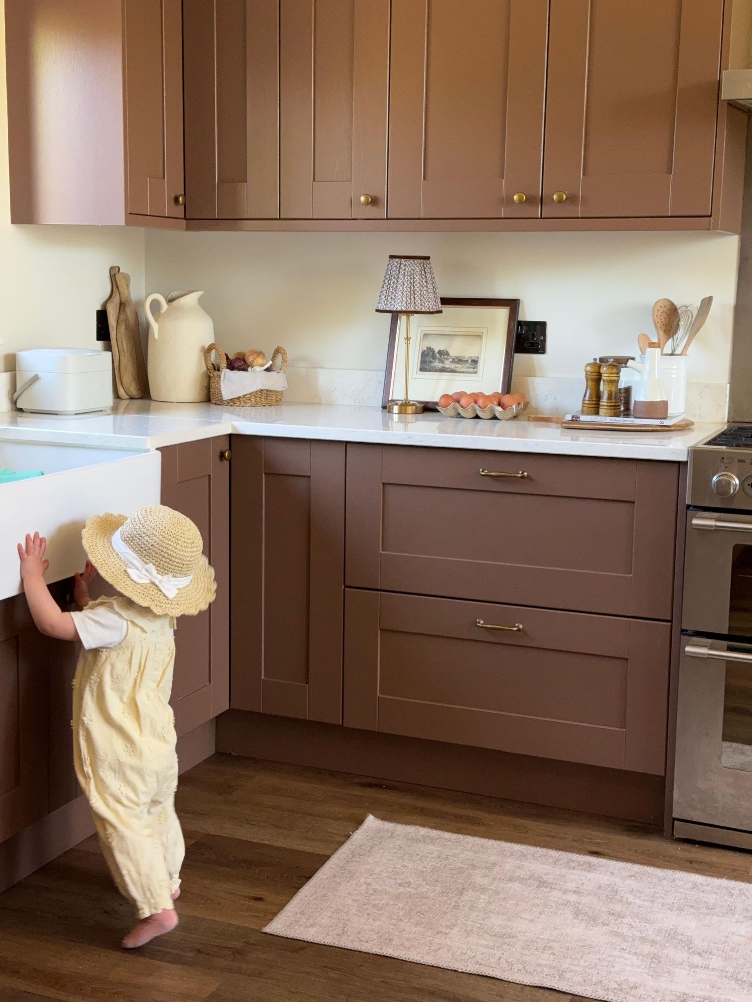 Toddler in yellow dress and straw hat stands in a modern kitchen with mauve cabinets and white marble countertops.