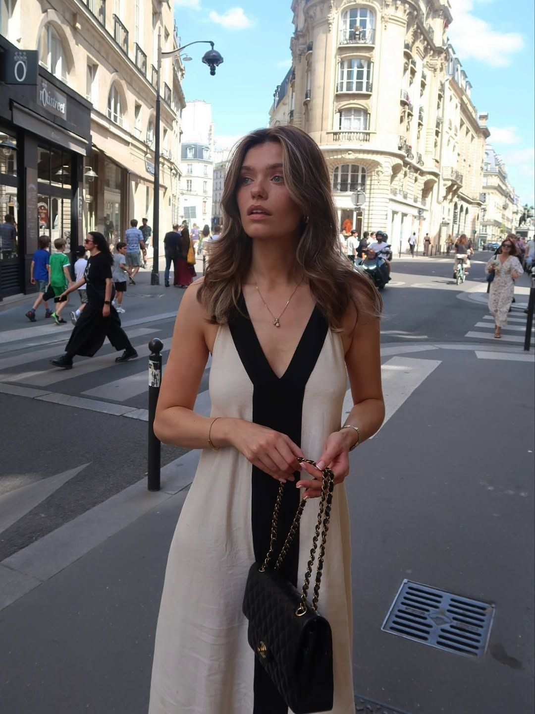 Woman in elegant black and cream maxi dress holding Chanel bag on a sunny Parisian street.