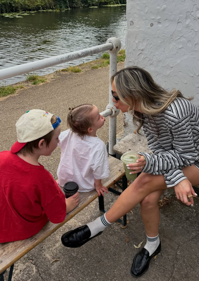 A woman shares a sweet moment with two young children on a bench by a river, holding iced drinks outdoors.