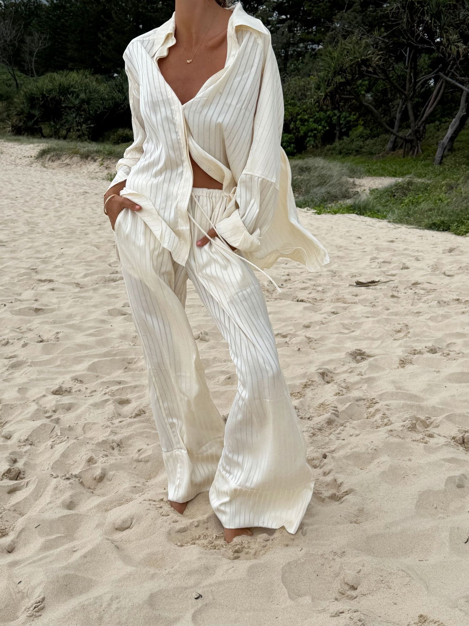 Woman wearing cream striped linen wide-leg pants and matching tie-front shirt on sandy beach.