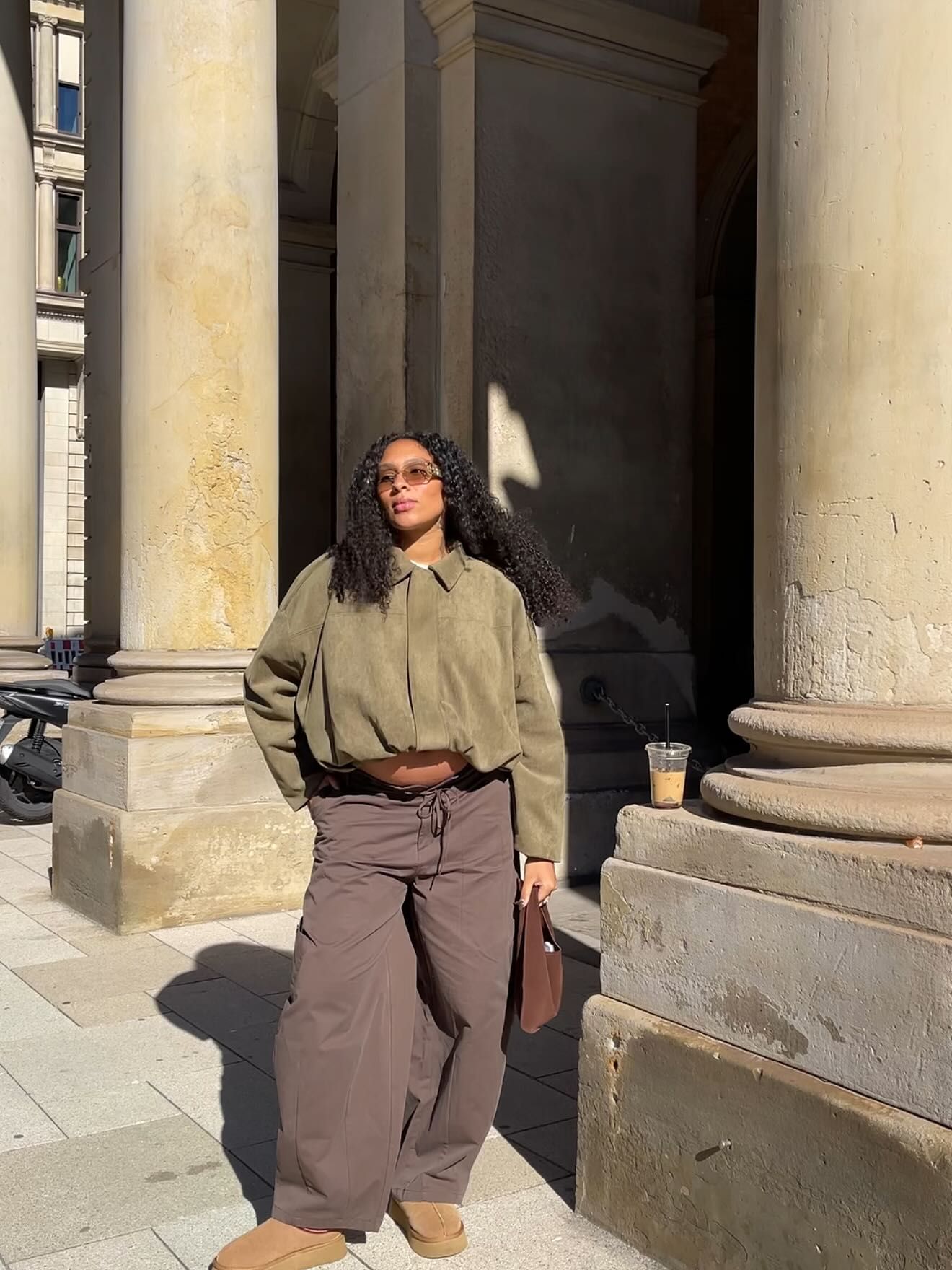 Woman in olive cropped jacket, brown wide-leg pants, and UGG boots poses by stone columns outdoors.