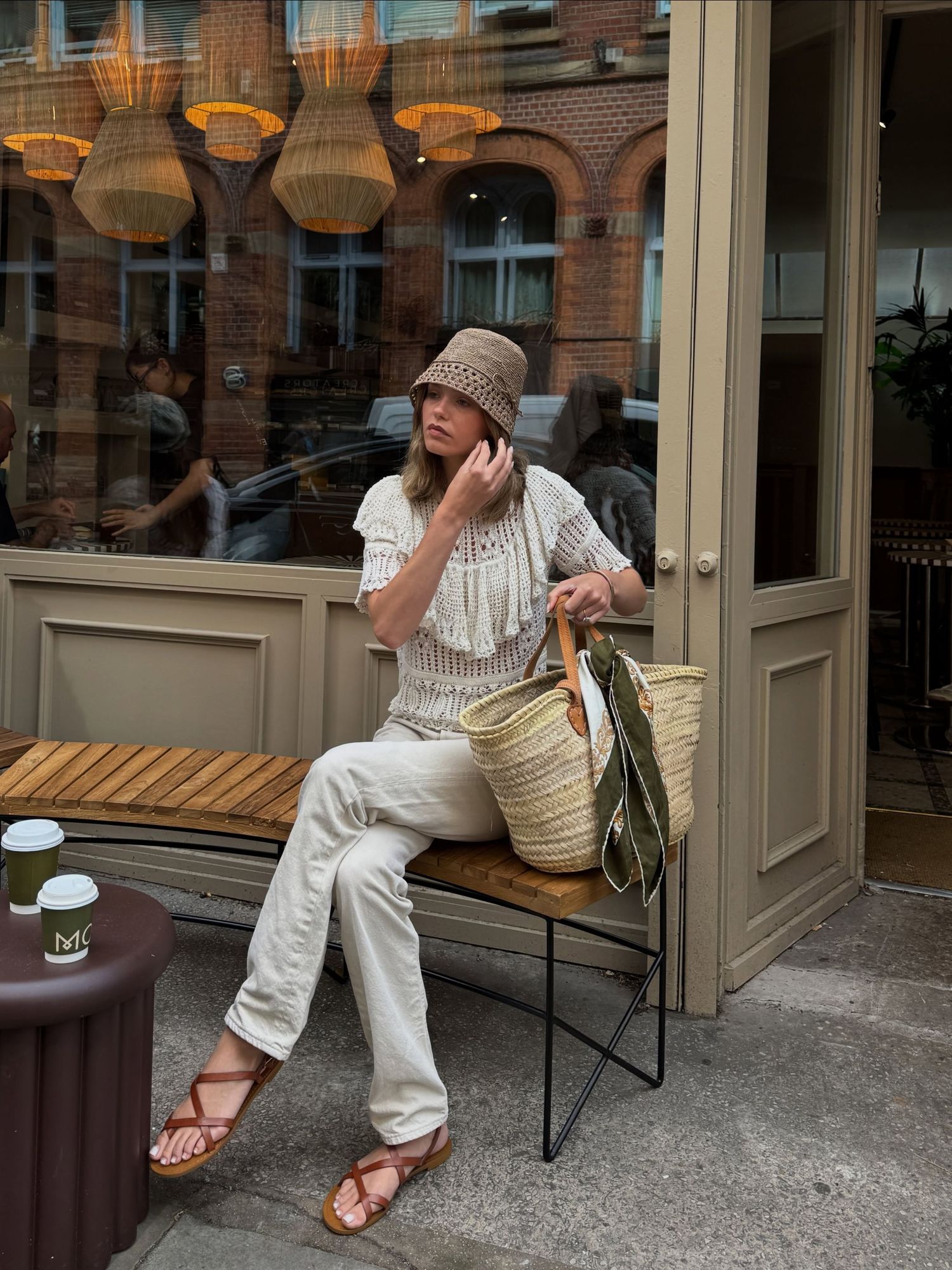 Woman in bucket hat, fringe top, white pants, and sandals sitting outside a cafe with a straw tote bag.