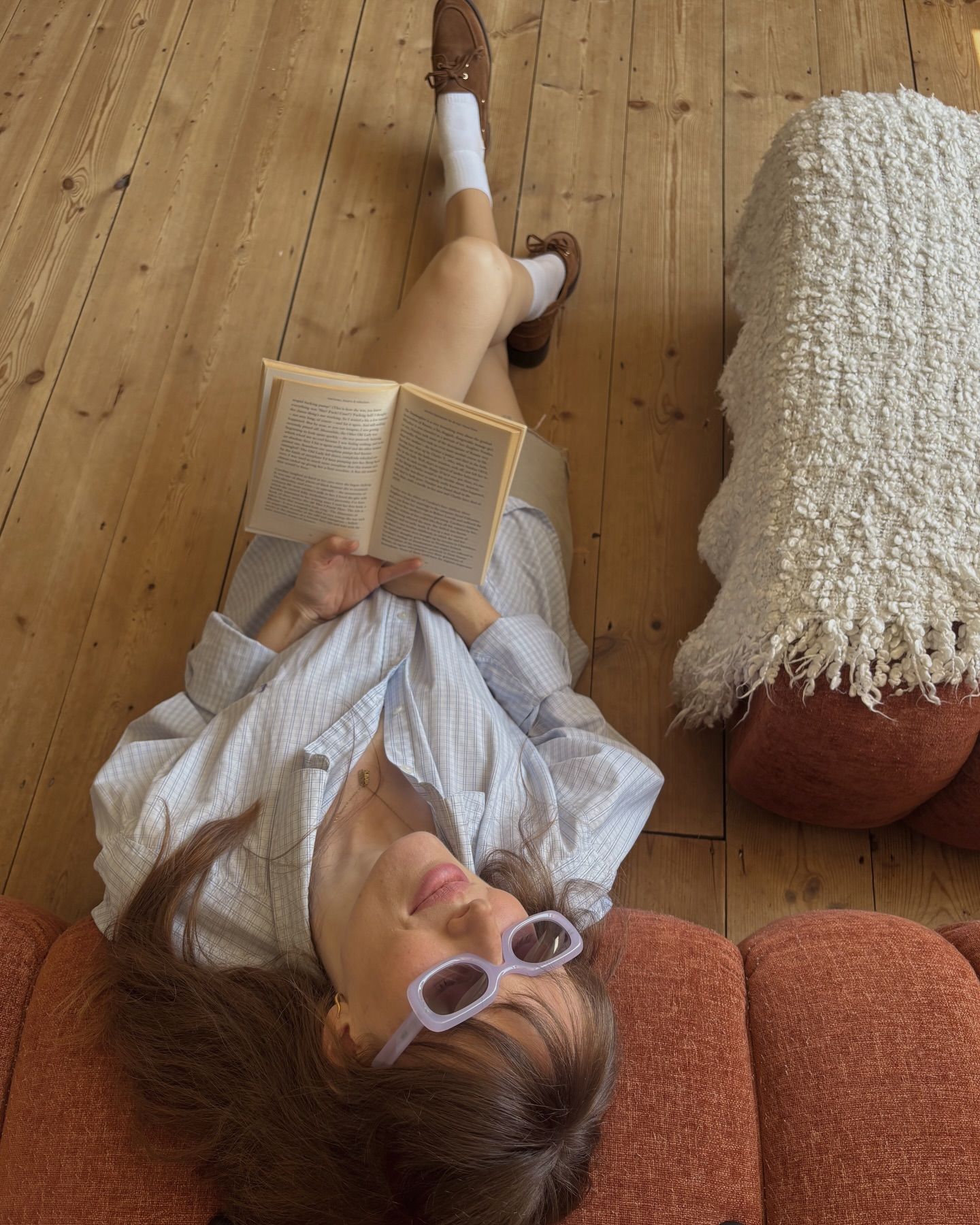 Woman relaxing on floor reading a book, wearing lavender sunglasses, white socks, and brown loafers on hardwood floors.