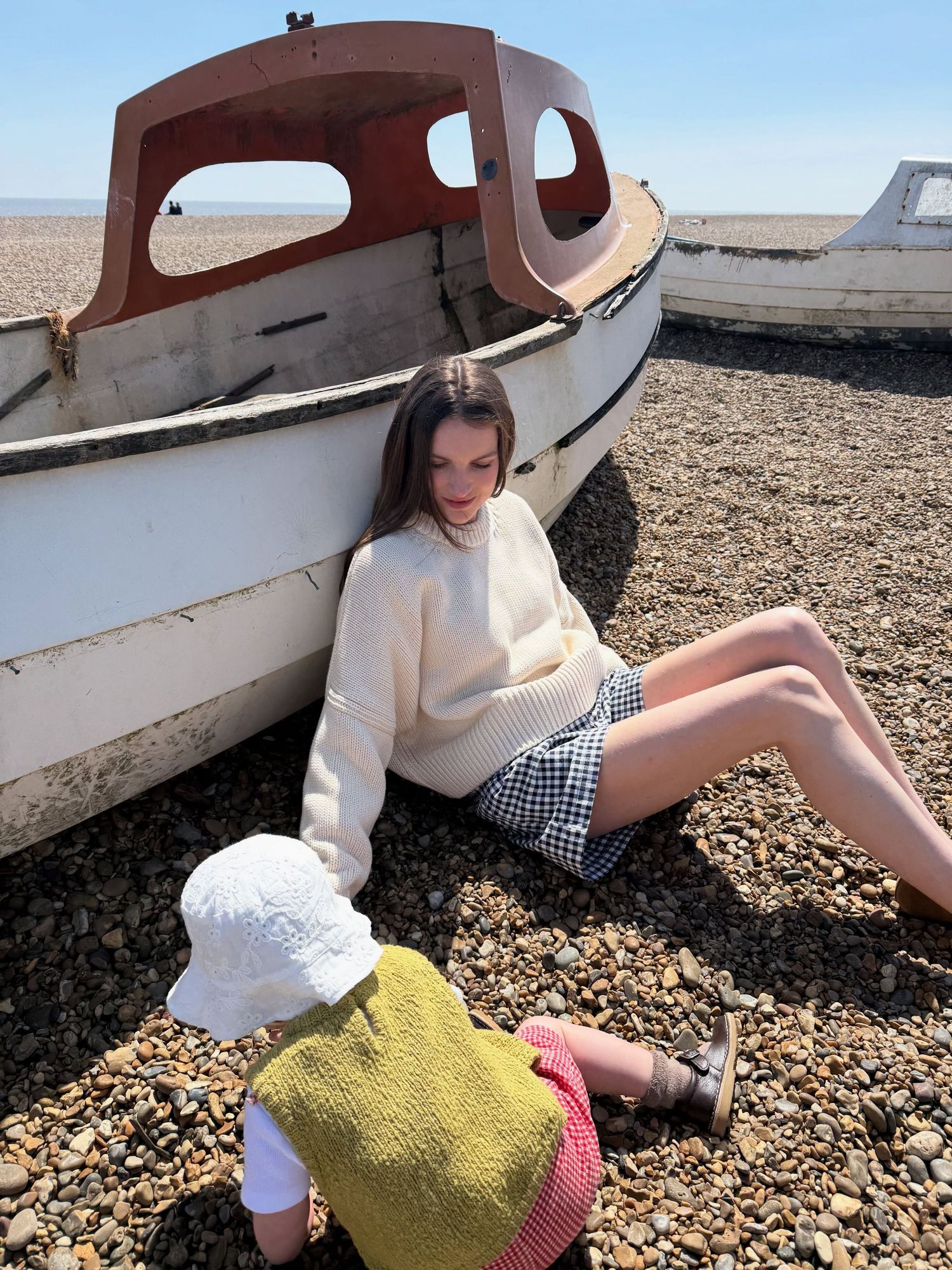 Woman in cream sweater and checkered shorts sits by a white boat on a pebble beach with a child in a yellow top.