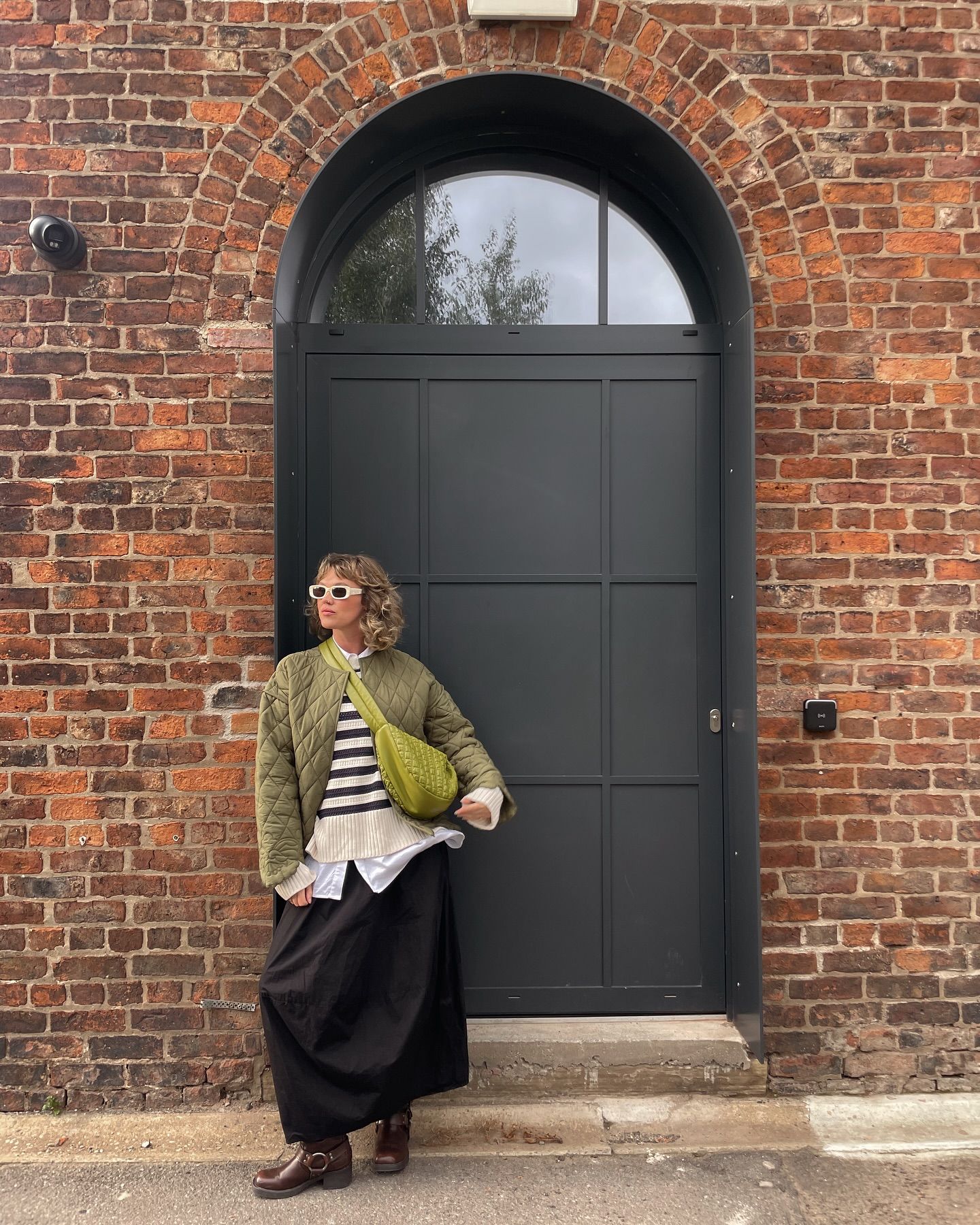 Person in striped top, green quilted jacket, black maxi skirt, posing by a dark arched door in a red brick wall.