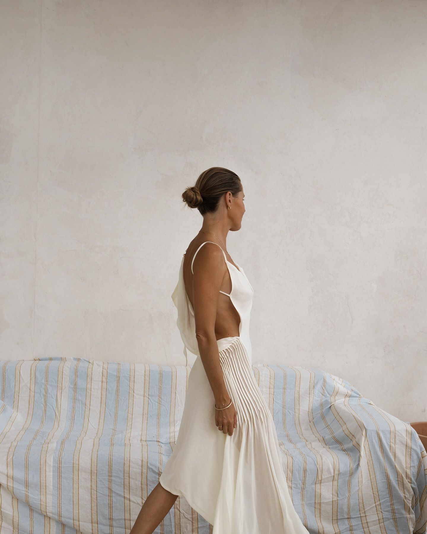 Woman in white sleeveless midi dress with open back walks beside a blue striped sofa against a neutral wall.
