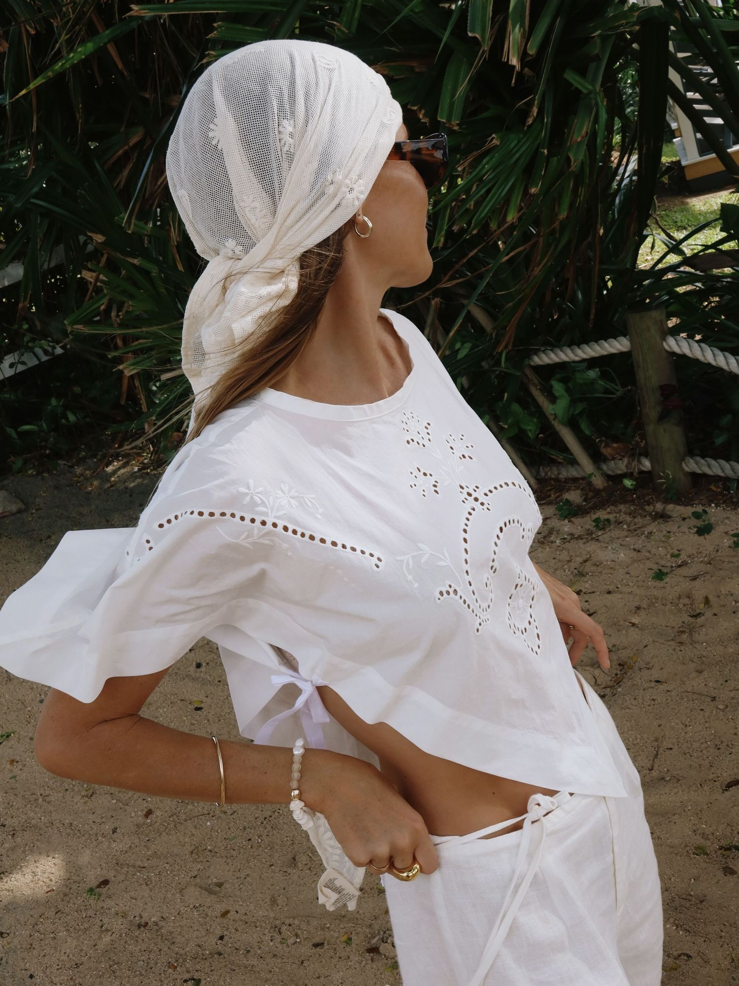 Woman in white eyelet crop top and matching skirt with white head scarf, posing outdoors near tropical greenery.